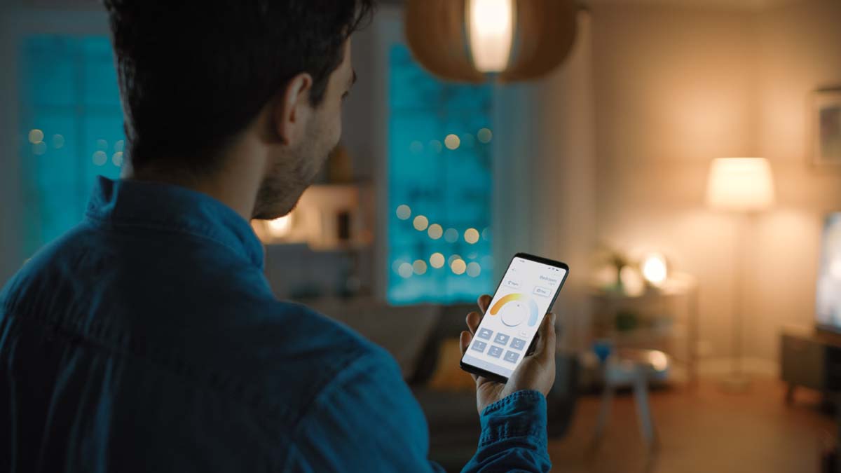 Young Handsome Man is Setting up Comfortable Bedroom Conditions with Voice Command to Smart Home Application on His Smartphone. It’s a Cozy Evening in Technologically Advanced Living Room.